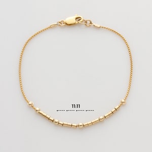 May include: A gold chain bracelet with small gold beads and cylinder-shaped beads. The bracelet has a clasp closure. The text "11:11" is printed below the bracelet.
