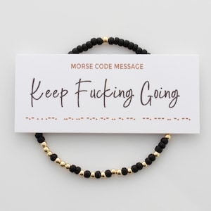 Könnte beinhalten: Schwarzes Perlenarmband mit goldenen Perlen und einer Karte mit der Aufschrift "Morse Code Message Keep Fucking Going" mit dem Morsecode für die Nachricht darunter.
