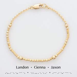 Könnte beinhalten: Ein goldenes Perlenarmband mit einem Verschluss. Das Armband hat ein schlichtes Design mit abwechselnd runden und rechteckigen Perlen. Der Text "Landon. Cienna. Jaxon" ist unter dem Armband gedruckt.