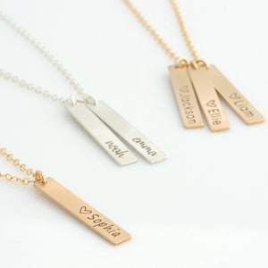 May include: Three gold bar necklaces with personalized names. The necklaces feature the names "Liam", "Ellie", and "Jackson" with a heart symbol before each name. A silver bar necklace with the names "Emma" and "Noah" is also shown.