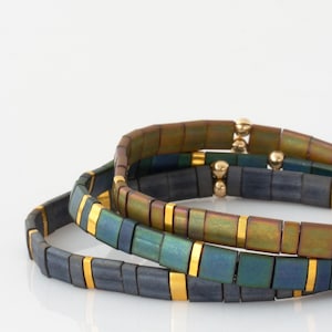 May include: Three stacked bracelets with a rectangular tile design. The tiles have a dark, iridescent finish with gold-colored accents. The bracelets are on a white background, with a slight reflection.