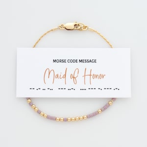 May include: A gold bracelet with purple beads and a clasp. A white card with the text "Morse Code Message Maid of Honor" and the morse code for the phrase is below the bracelet.