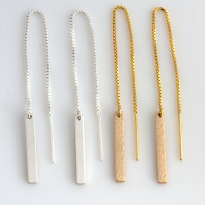 Puede incluir: Cuatro pares de pendientes colgantes. Dos pares son plateados, con barras rectangulares y cadena. Los otros dos pares son dorados, con barras rectangulares texturizadas y cadena. Los pendientes se muestran sobre un fondo blanco.