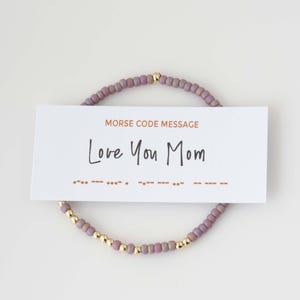 May include: A beaded bracelet with a Morse code message card. The bracelet features small, mauve-colored beads interspersed with gold-colored beads. The card reads "Love You Mom" in black script, with Morse code symbols below.