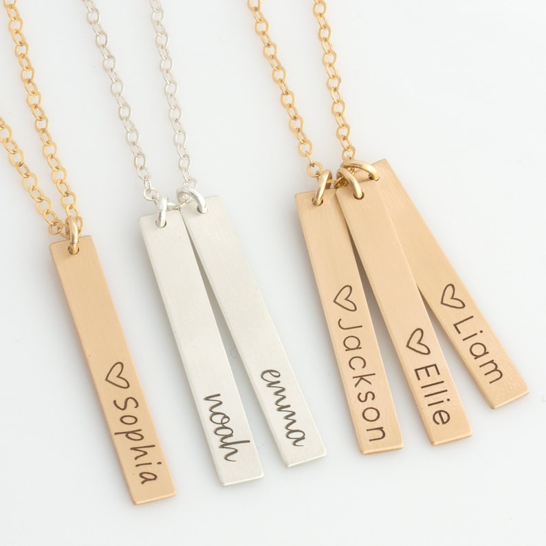 May include: Four gold and silver bar necklaces with personalized names, including "Sophia", "Noah", "Emma", "Jackson", "Ellie", and "Liam". Each bar has a small heart symbol above the name.