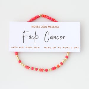 May include: A beaded bracelet with red and gold beads, encircling a white card. The card reads "MORSE CODE MESSAGE" above the words "Fuck Cancer" in a handwritten font. Below the text is a line of Morse code.