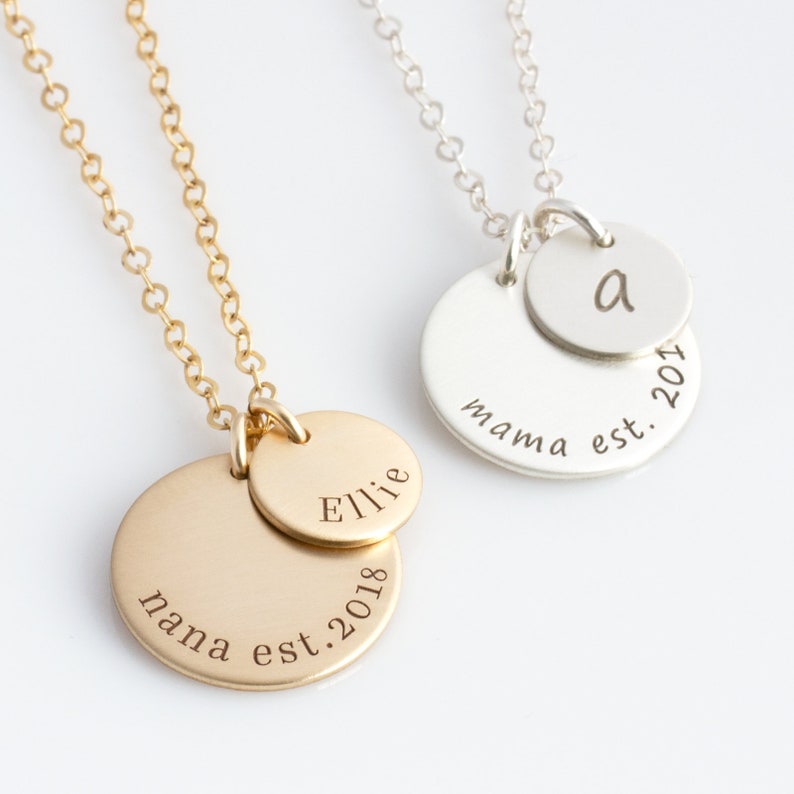 May include: Two gold and silver necklaces with personalised engraved discs. The gold necklace has a larger disc engraved with "nana est. 2018" and a smaller disc engraved with "Ellie". The silver necklace has a larger disc engraved with "mama est. 2017" and a smaller disc engraved with the letter "a".