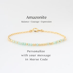 May include: A delicate gold bracelet featuring small, pale green amazonite beads and gold accents. The bracelet is labeled "Amazonite" with the words "Balance, Courage, Expression" and "Personalize with your message in Morse Code".