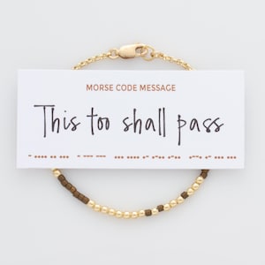 Könnte beinhalten: Gold- und braunes Perlenarmband mit einer Karte, auf der in Morsecode "This too shall pass" steht.
