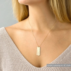 May include: Gold necklace with three vertical bar pendants, each engraved with a name. The names are "Ella", "Jackson", and "Olivia". The model is wearing an 18-inch chain.