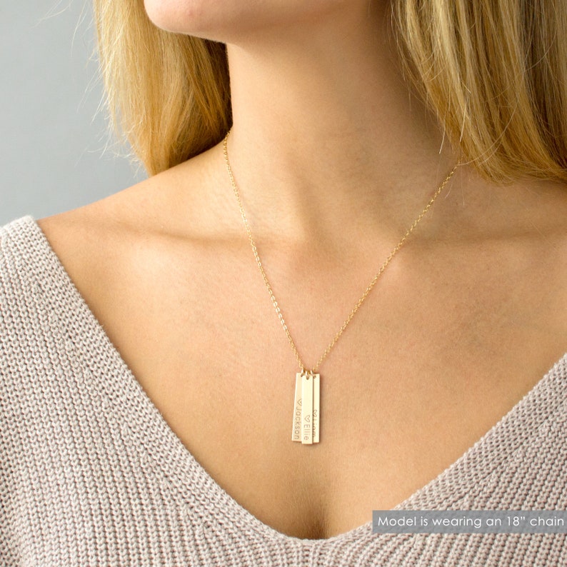 May include: Gold necklace with three vertical bar pendants, each engraved with a name. The names are "Ella", "Jackson", and "Olivia". The model is wearing an 18-inch chain.