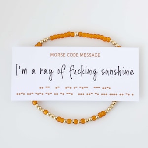 May include: A beaded bracelet with amber-colored beads and gold-colored accents encircles a white card. The card reads "I'm a ray of fucking sunshine" in cursive, with a Morse code message below. The words "MORSE CODE MESSAGE" are printed above the text.