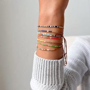 Könnte beinhalten: Eine Sammlung farbenfroher Perlenarmbänder, die an einem Handgelenk getragen werden. Die Armbänder weisen verschiedene Farbkombinationen auf, darunter Schwarz und Weiß, Blau, Rot und Grün. Die Armbänder sind auf dünnen Schnüren aufgefädelt und mit einem braunen Faden gebunden.