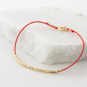 May include: A red string bracelet with gold beads. The bracelet has a gold clasp.