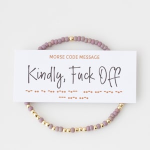 May include: A beaded bracelet with mauve-colored beads and gold-colored accent beads. A white card with the text "MORSE CODE MESSAGE" and "Kindly, Fuck Off" is placed over the bracelet.