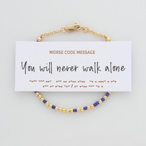 May include: A gold chain bracelet featuring alternating small gold beads and blue beads, displayed with a white card that reads MORSE CODE MESSAGE, You will never walk alone, and includes the corresponding Morse code dot and dash sequence.