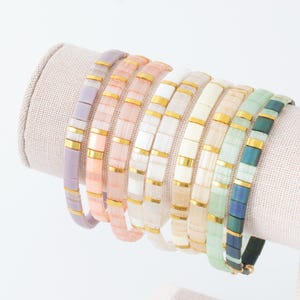 May include: A collection of eight stretch bracelets arranged in a row, each featuring rectangular beads in various pastel and vibrant colors including lavender, peach, pink, white, cream, beige, light green, and dark blue, accented with gold spacers.