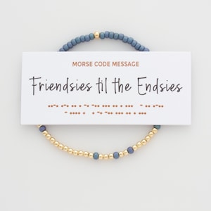 May include: A friendship bracelet with gold and blue beads, and a card that reads "Friendsies til the Endsies" in script. The card also has a Morse code message. The bracelet is on a white background.