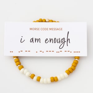 Könnte beinhalten: Ein Armband mit senfgelben und weißen Perlen und goldenen Akzenten. Eine Karte über dem Armband zeigt "i am enough" in Schreibschrift, wobei die Nachricht auch in Morsecode angezeigt wird.
