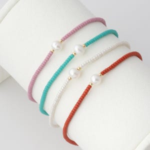 May include: Four beaded bracelets in pink, turquoise, white, and red. Each bracelet features a single white pearl and gold-colored accents. The bracelets are displayed on a white, curved surface, showcasing their delicate design and color variations.