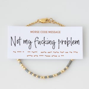Könnte beinhalten: Ein goldfarbenes Armband mit Kette und Verschluss, kombiniert mit einer weißen Karte. Auf der Karte steht in schwarzer Schrift "Not my fucking problem", darunter eine Morsecode-Nachricht. Das Armband hat goldene und graue Perlen.
