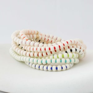 May include: A stack of multiple beaded bracelets featuring white, round beads interspersed with small, colorful accent beads in shades of red, black, light green, turquoise, and blue, along with small gold-toned metallic spacer beads.
