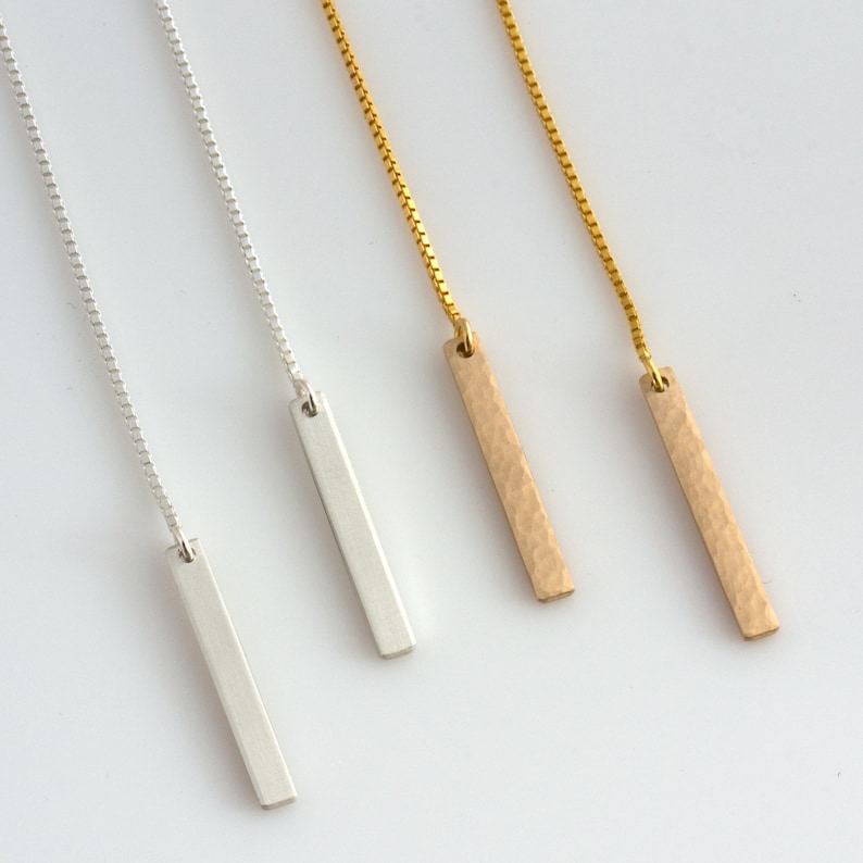 Puede incluir: Cuatro collares minimalistas con barras. Dos son plateados, con un acabado liso, y dos son dorados, con una textura martillada. Cada collar tiene una delicada cadena.