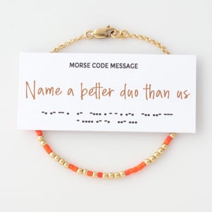 May include: A gold chain bracelet with orange and gold beads. The bracelet has a card attached to it that says "Name a better duo than us" in Morse code.