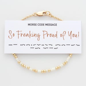 May include: A gold chain bracelet with white beads. The bracelet has a card attached to it that says "So Freaking Proud of You!" in brown text and the message in Morse code.