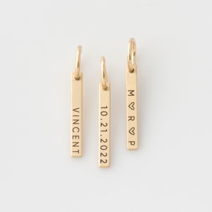 Peut inclure: Trois pendentifs en forme de barre dorés avec des gravures différentes. Le premier pendentif dit "Vincent", le deuxième pendentif dit "10.21.2022" et le troisième pendentif dit "M♥R♥P".