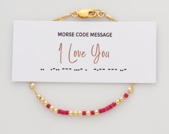 Könnte beinhalten: Ein goldenes Kettenarmband mit abwechselnd kleinen goldenen Perlen und rosafarbenen zylindrischen Perlen. Eine weiße Karte ist befestigt, die den Text MORSE CODE MESSAGE, I Love You und eine Morsecode-Sequenz in schwarzer Tinte zeigt.