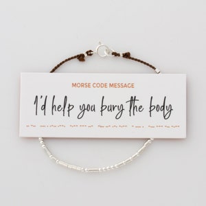 Könnte beinhalten: Ein braunes Kordelarmband mit silbernen Perlen, die in Morsecode "Ich würde dir helfen, den Körper zu begraben" buchstabieren. Das Armband wird mit einer Karte geliefert, die die Übersetzung in Morsecode zeigt.