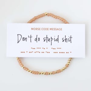 May include: A beaded bracelet featuring alternating light tan and metallic gold beads. A white rectangular card rests on top, displaying the text MORSE CODE MESSAGE, the phrase Don't do stupid shit, and the corresponding Morse code sequence in dots.