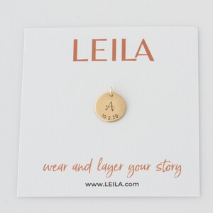 May include: Gold disc pendant with the letter "A" and the date "10.2.20" engraved on it. The pendant is on a white card with the word "LEILA" printed in red at the top and the text "wear and layer your story www.LEILA.com" at the bottom.