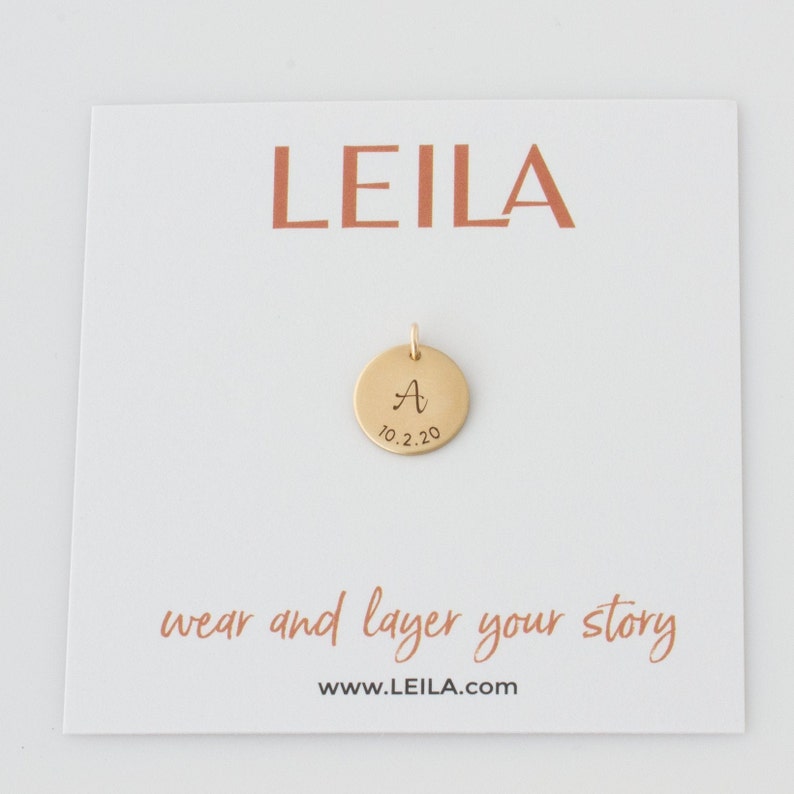 May include: Gold disc pendant with the letter "A" and the date "10.2.20" engraved on it. The pendant is on a white card with the word "LEILA" printed in red at the top and the text "wear and layer your story www.LEILA.com" at the bottom.