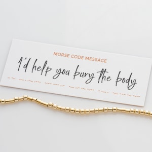 May include: A white card with the text "Morse Code Message" and the phrase "I'd help you bury the body" written in black cursive. A gold beaded bracelet is lying on the card.