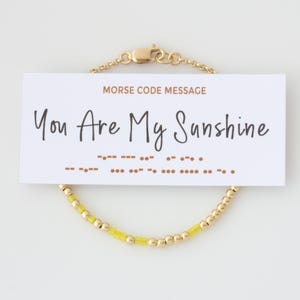 May include: A gold-colored bracelet with yellow beads and a Morse code message card. The card reads "You Are My Sunshine" in black script. The bracelet has a delicate chain and clasp.
