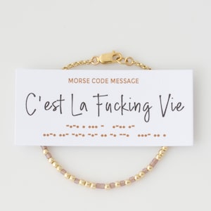May include: A gold-toned bracelet with a chain and clasp, featuring a strand of small gold beads and mauve-colored square beads. A white card with the text "C'est La Fucking Vie" and Morse code is attached.