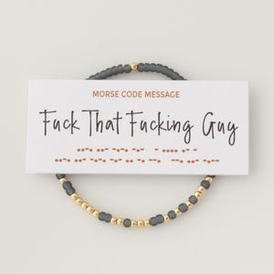 May include: A beaded bracelet with a card that reads "Fuck That Fucking Guy." The bracelet features gray and gold beads. The card also includes a Morse code message.