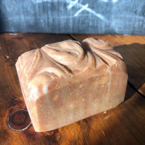 May include: A rectangular bar of handmade soap with a swirled, textured design on top. The soap has a light brown color with flecks of darker brown and white. The soap is sitting on a wooden surface.