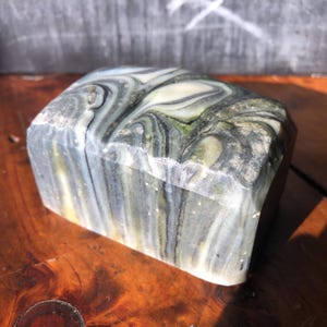 May include: A rectangular, hand-carved stone soap dish with a unique, swirled pattern of gray, green, and cream colors. The soap dish has a faceted design and a polished finish, showcasing the natural stone's beauty.