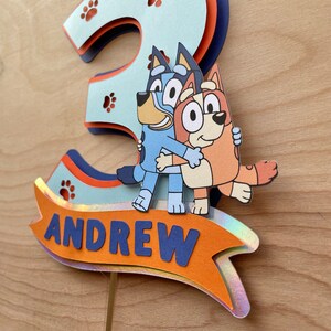 Puede incluir: Un adorno para pastel azul y naranja con los personajes Bluey y Bingo del programa de dibujos animados Bluey. El adorno tiene la forma del n&uacute;mero 3 y tiene el nombre "ANDREW" escrito en &eacute;l.
