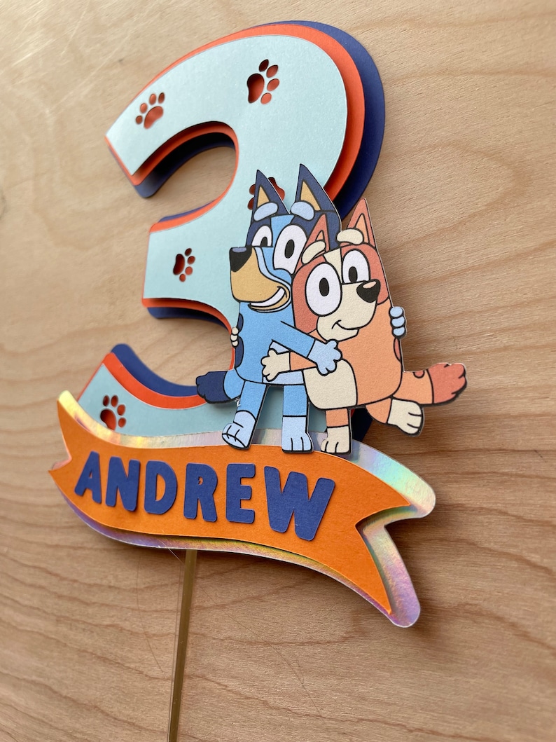 Puede incluir: Un adorno para pastel azul y naranja con los personajes Bluey y Bingo del programa de dibujos animados Bluey. El adorno tiene la forma del n&uacute;mero 3 y tiene el nombre "ANDREW" escrito en &eacute;l.