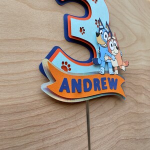 Puede incluir: Un topper de pastel en 3D de color azul y naranja con forma de n&uacute;mero tres, con huellas de patas y dos perros de dibujos animados. El topper tiene una pancarta que dice "ANDREW".