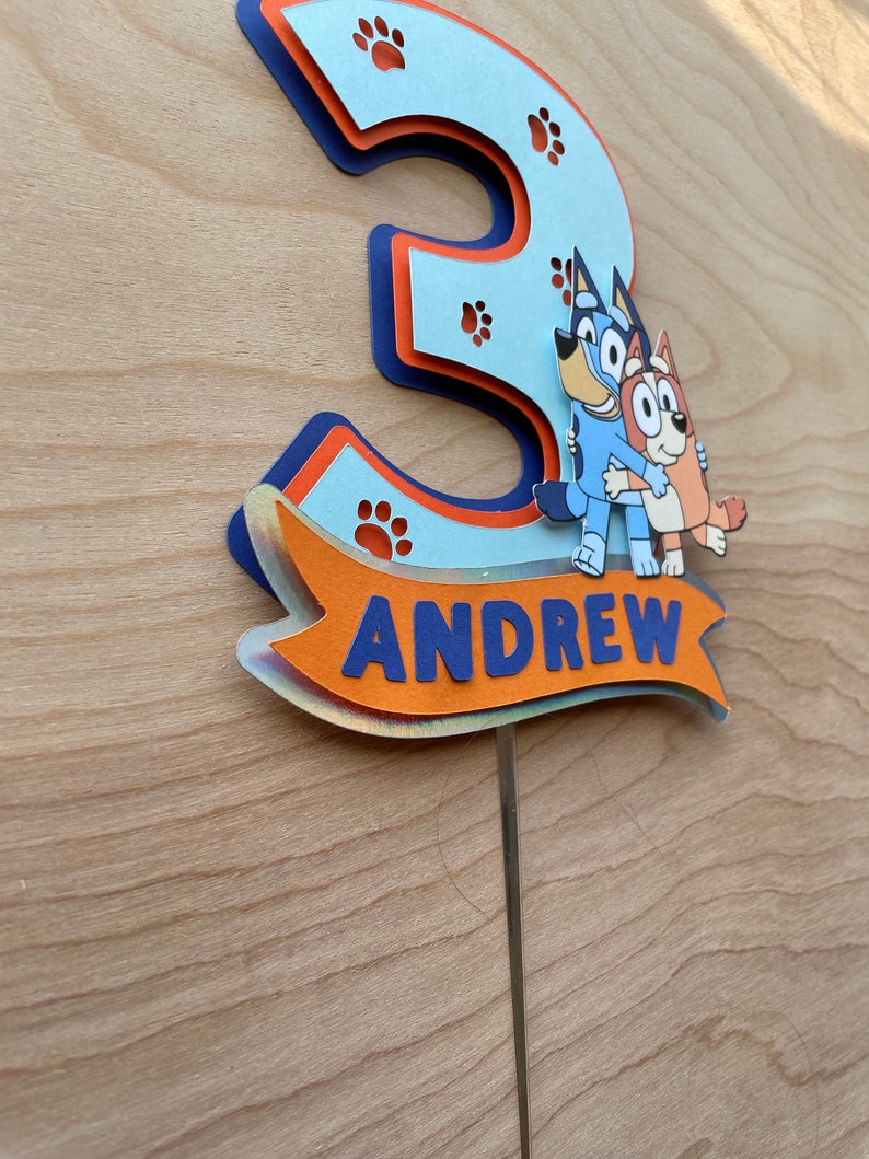 Puede incluir: Un topper de pastel en 3D de color azul y naranja con forma de n&uacute;mero tres, con huellas de patas y dos perros de dibujos animados. El topper tiene una pancarta que dice "ANDREW".