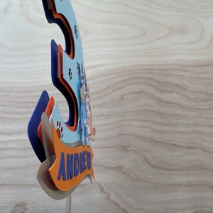 Puede incluir: Un adorno para pastel 3D azul y naranja con un perro de dibujos animados y el nombre "ANDREW" en una pancarta. El adorno est&aacute; en un palo de pl&aacute;stico transparente.