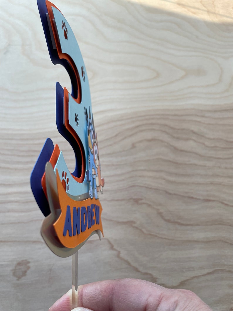Puede incluir: Un adorno para pastel 3D azul y naranja con un perro de dibujos animados y el nombre "ANDREW" en una pancarta. El adorno est&aacute; en un palo de pl&aacute;stico transparente.