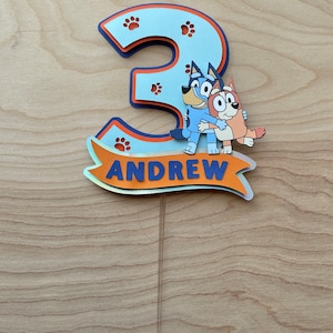 Puede incluir: Un adorno para pastel azul y naranja con el n&uacute;mero 3, huellas de patas y el texto "ANDREW". Dos perros de dibujos animados se abrazan frente al n&uacute;mero.