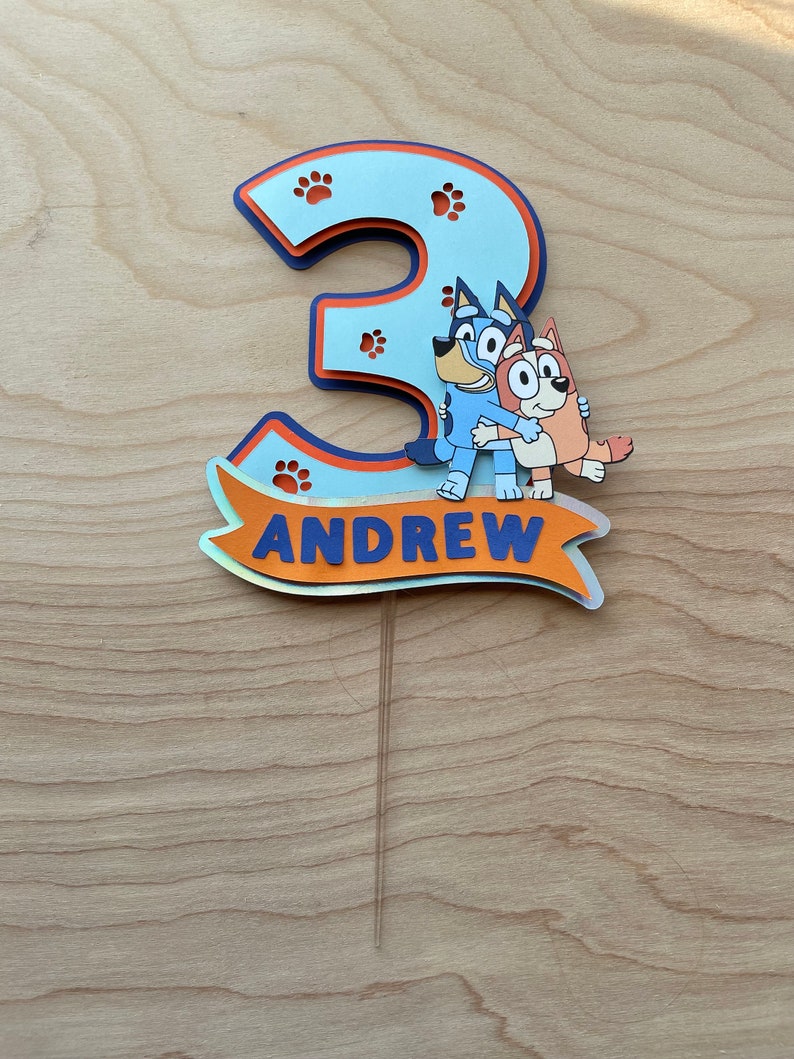 Puede incluir: Un adorno para pastel azul y naranja con el n&uacute;mero 3, huellas de patas y el texto "ANDREW". Dos perros de dibujos animados se abrazan frente al n&uacute;mero.