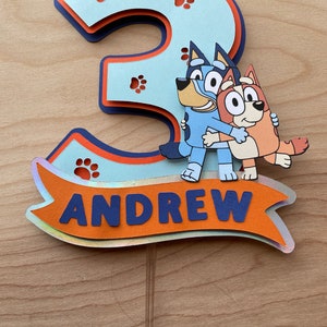 Puede incluir: Un adorno para pastel 3D azul y naranja con el n&uacute;mero 3, huellas de patas y los personajes del programa Bluey. El adorno tambi&eacute;n incluye el nombre Andrew en una pancarta.